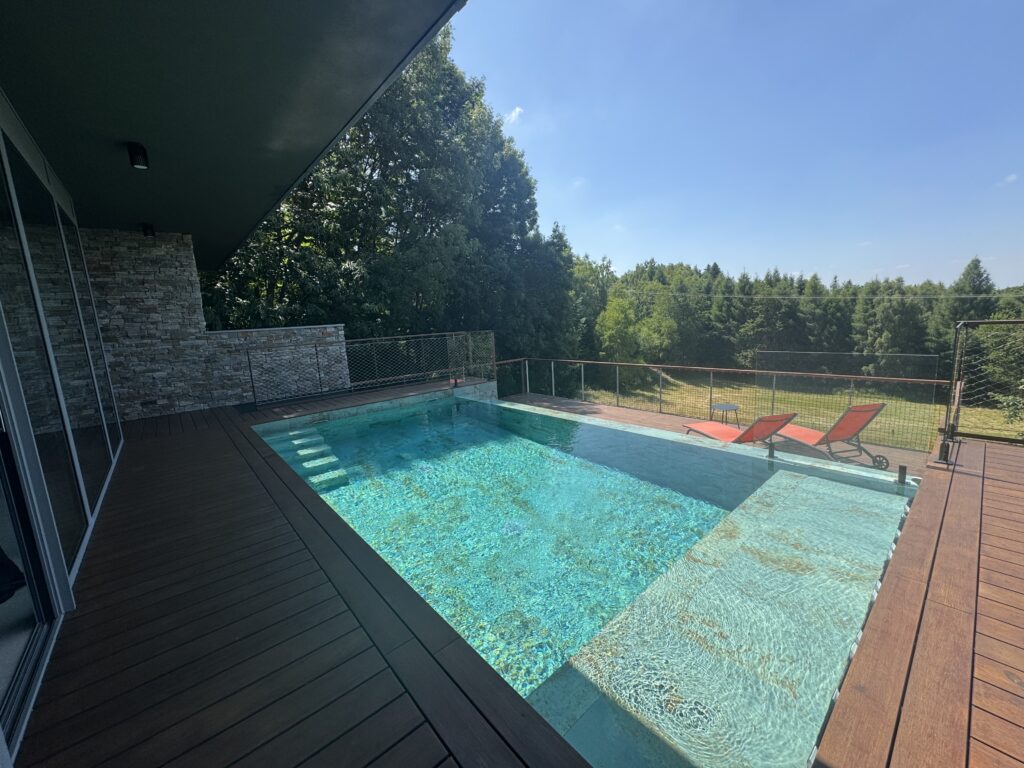 IMG_2077 - Villa Concept 57 - Villa Concept 57 Outdoor pool area with a wooden deck, red lounge chairs, and glass railing, adjacent to a stone-clad wall and a covered patio, with trees and a clear blue sky in the background.
