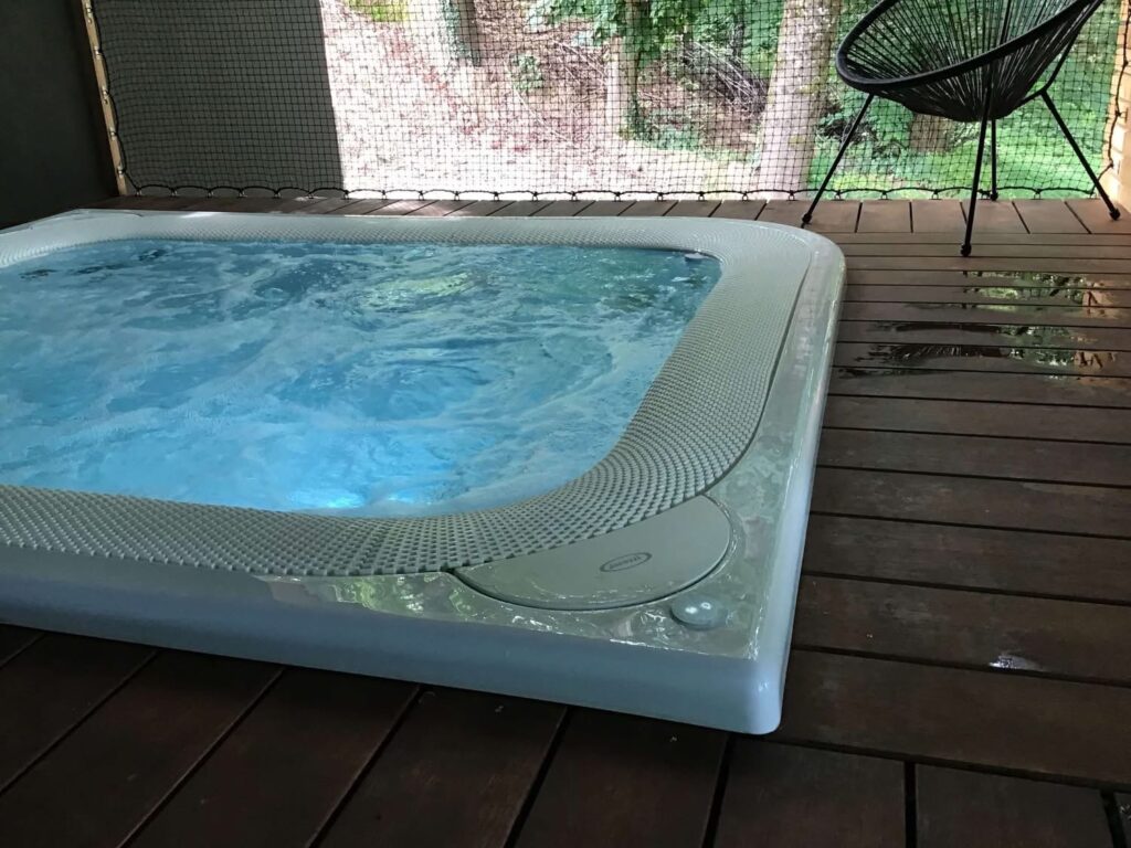 Outdoor hot tub on a wooden deck with a netted barrier and a black chair in the background; water ripples visible in the tub.
